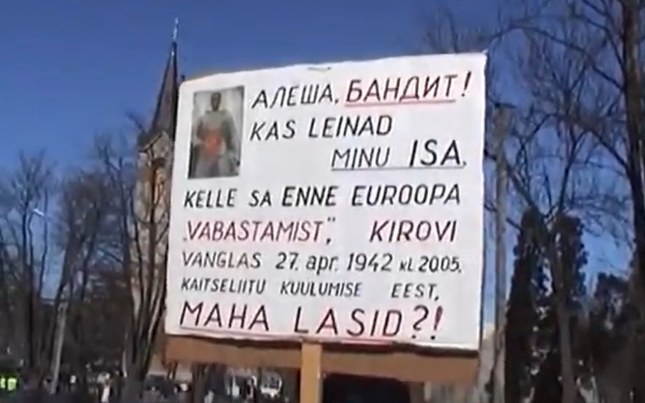 К Бёму присоединяется пожилой седовласый мужчина. Он берёт в руки плакат, тогда как флаг остаётся у Бёма. Этот жест становится поворотным. Уже по дороге к памятнику провокация начинает работать: в адрес старика раздаётся грубое оскорбление по-русски. Оно адресовано конкретному человеку с конкретным плакатом. Не флагу. Не государству. Не народу. Но камера не задаёт вопросов — она просто фиксирует звук, вырывая его из контекста.