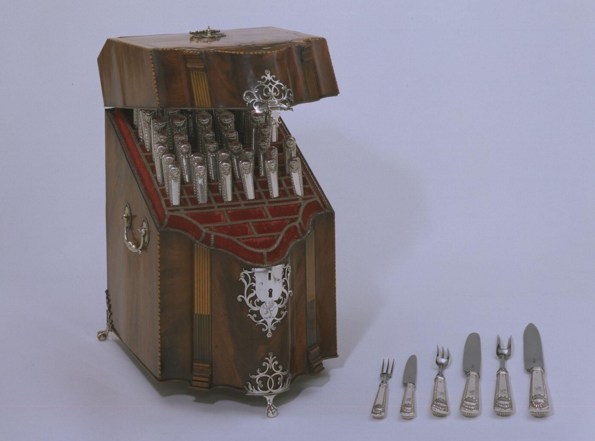 Коробка для ножей (Knife box). Между 1775-1785. Шпон красного дерева с серебряной фурнитурой. Размеры: высота: 38 см, ширина: 23 см, глубина: 34,5 см. Виктория и Альберт музей, Лондон.                      Подобные коробки для ножей были очень популярны в Англии во второй половине XVIII века. Их обычно ставили парами по обе стороны буфета или серванта, а внутри хранили не только ножи, но и вилки, а иногда и приборы для разделки мяса. Позднее именно из таких коробок эволюционировали более вместительные кабинеты и шкафчики для хранения столовых приборов XIX века.
