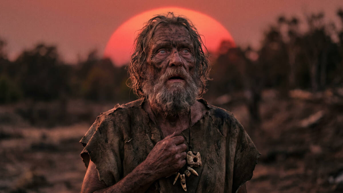 На фотографии изображен пожилой бородатый мужчина в рваной грязной одежде с костяным амулетом, смотрящий вверх на фоне большого красного закатного солнца и силуэтов деревьев. Изображение сгенерировано в ИИ для иллюстрации идеи.