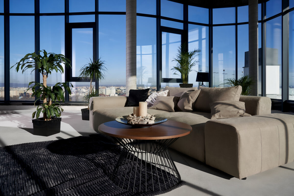    Front view of fashionable interior spacious room with large panoramic window with incredible view on city and cozy grey sofa with pillows and green flowerpots. Concept of modern interior. Фелор Круглов