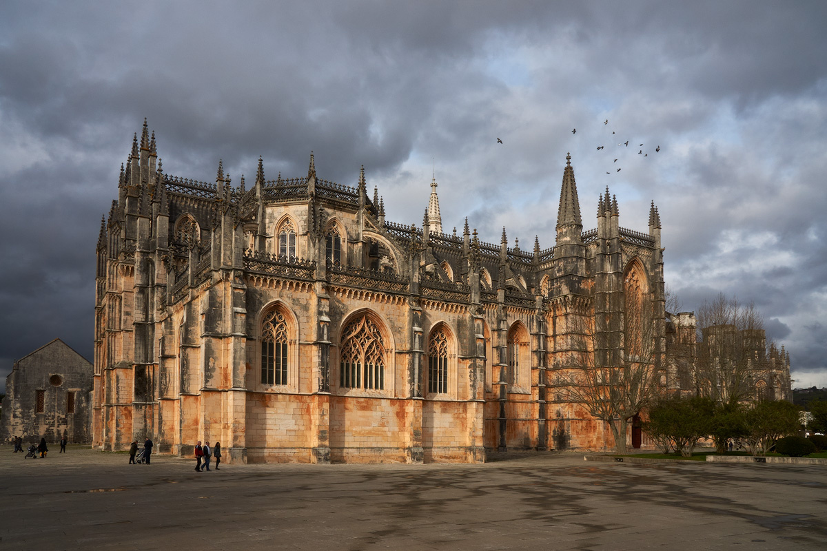 Монастырь Баталья (Mosteiro da Batalha) — не просто архитектурный памятник, а каменная летопись португальской независимости. Ниже — подробный разбор с конкретными фактами, цифрами и малоизвестными деталями.
---
Точные даты и хронология строительства
* 1385 год — битва при Алжубарроте: португальцы под командованием короля Жуана I и полководца Нуну Алвареша Перейры разгромили кастильцев, обеспечив независимость страны.
* 1386 год — начало строительства монастыря по обету короля: он пообещал возвести обитель в честь Девы Марии, если одержит победу.
* Около 1517 года — официальное прекращение масштабных работ: ресурсы и лучшие мастера были перенаправлены на строительство монастыря Жеронимуш в Лиссабоне.
* Таким образом, строительство длилось более 130 лет, и в нём участвовали не менее 15 архитекторов, каждый из которых вносил свои изменения.
---
Архитектурные факты и цифры
* Высота нефа достигает примерно 32 метров — это создаёт ощущение устремлённости вверх и подчёркивает готический характер здания.
* Западный фасад украшен статуями: здесь можно увидеть фигуры ветхозаветных царей, пророков, апостолов и ангелов, каждая из которых стоит под отдельным каменным балдахином — редкий пример такой детальной проработки в португальской архитектуре.
* Витражи относятся к концу XV — началу XVI века и считаются одними из старейших в Португалии. Они изображают сцены из жизни Христа и Девы Марии и до сих пор сохраняют часть оригинальной палитры.
* Южный портал — крупнейший готический портал в Португалии: его окно — одно из самых больших в национальной готике, а резьба включает гербы и символику династии Авис.