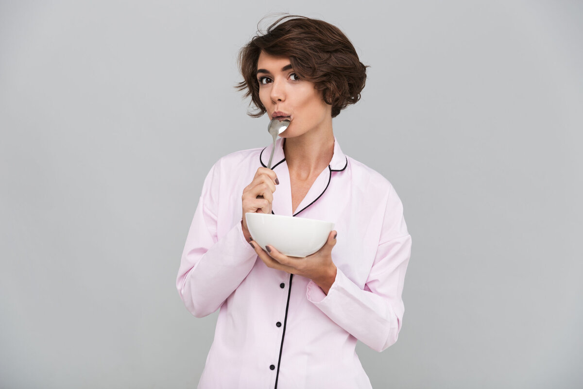    Portrait of an attractive young girl in pajamas holding a bowl with cereal and licking a spoon isolated over gray background