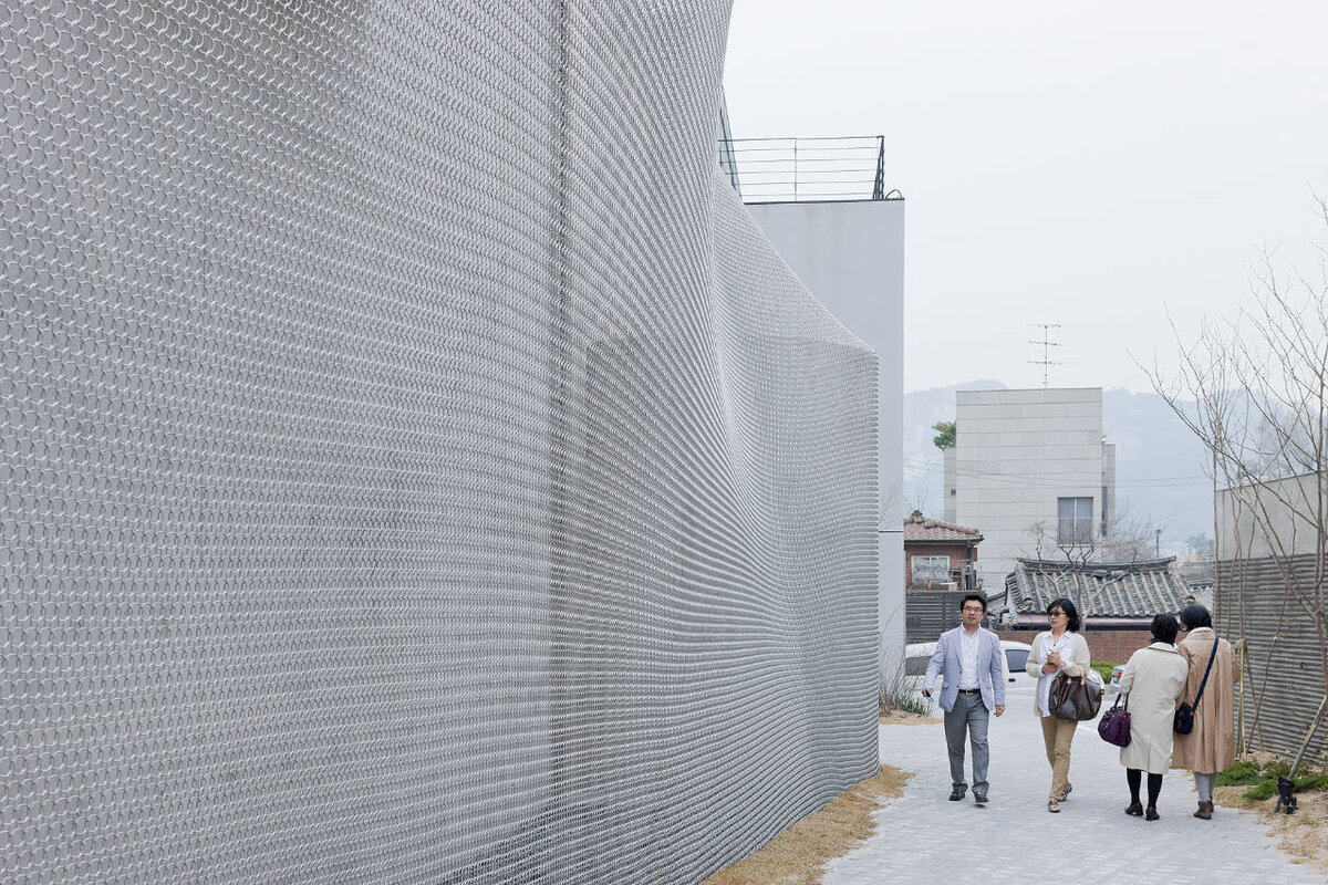 Изготовленная на заказ сетка из нержавеющей стали создает слой диффузии вокруг конструкции благодаря сочетанию отражений, открытости и муаровых узоров, создаваемых за счет взаимодействия ее теней.