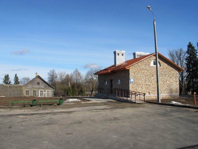 Итак, наше сооружение стоит на обочине шоссе, напротив своротка на Бор Бельково, на территории только что построенной стоянки для туристов. На виде со стороны стоянки заметно что сооружение облицовано как бы под местный известняк, имеет перекрытие под черепицу, две вытяжные трубы каминных размеров. В сооружении есть несколько полукруглых окон. Вход оборудован пандусом для инвалидов. На фоне окружающих изб туалет смотрится просто монстром.