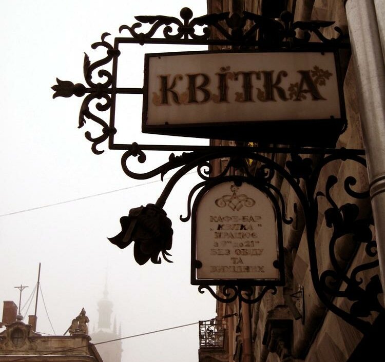 Дешевые материалы, конечно, развращают и отупляют. Раньше банерная печать не была доступна и хозяева лавок делали лаконичные вывески, которые отлично вписывались в окружающую архитектуру. Они были выпуклы и натуральны. Часто они делались перпендикулярно дороге, на специальных кронштейнах. Оно и понятно — пешеход потенциальный клиент и должен видеть вывеску прямо по пути своего движения. Выглядело это как-то так: 

