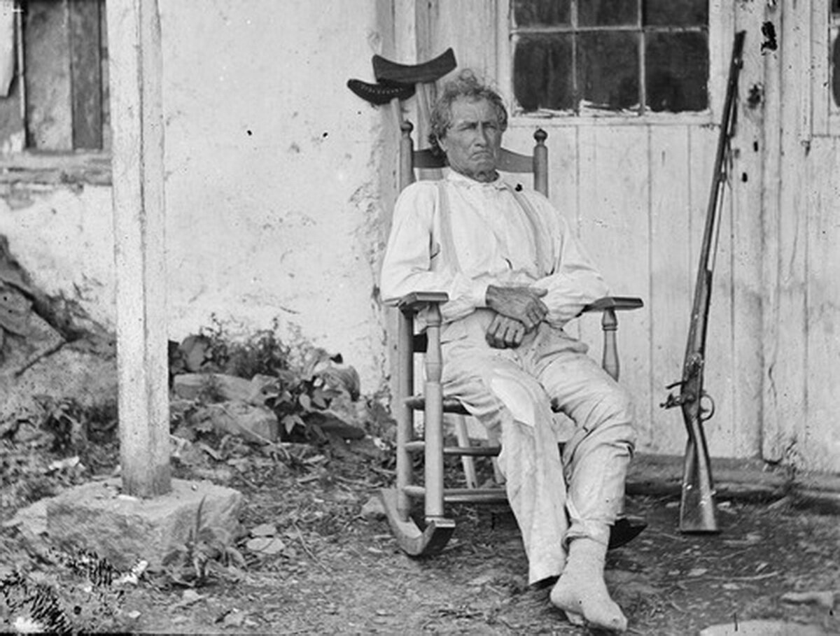 Джон Бёрнс (John Burns) сидит в кресле-​качалке после битвы при Геттисберге (Gettysburg). Это было самое кровопролитное сражение в ходе Гражданской войны в США, произошедшее 1-3 июля 1863 года в округе Адамс штата Пенсильвания и считающееся переломной точкой в конфликте. Ветеран войны 1812 года (имеется в виду англо-​американская война), 70-​летний Бёрнс взял винтовку и примкнул к армии Союза в качестве снайпера. Был ранен несколько раз.
