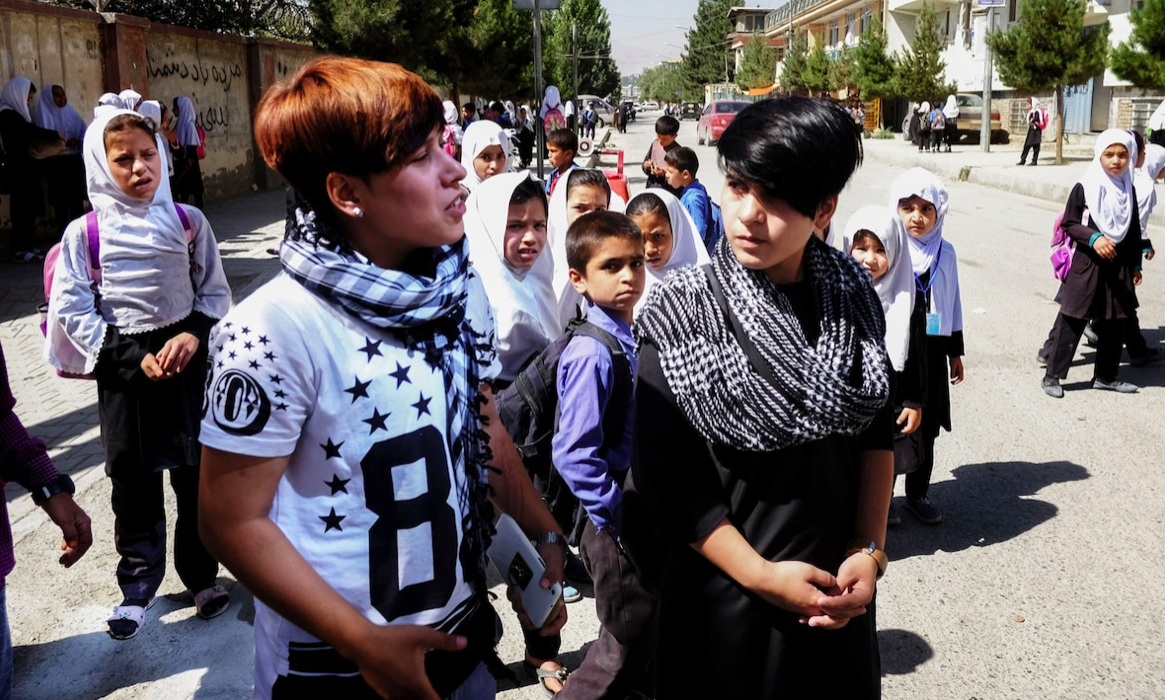 На фото: Сетар и Али среди детей, которые всячески комментируют их появление у школы. Не все относятся к "бача-пош" хорошо. "Люди подходят ко мне, спрашивают, почему я одеваюсь как мальчик", - рассказывает Сетар. Отец возит Али в школу, а Сетар перестала ходить туда, ей надоело, когда ее обзывают. Фото: Лулу д'Аки