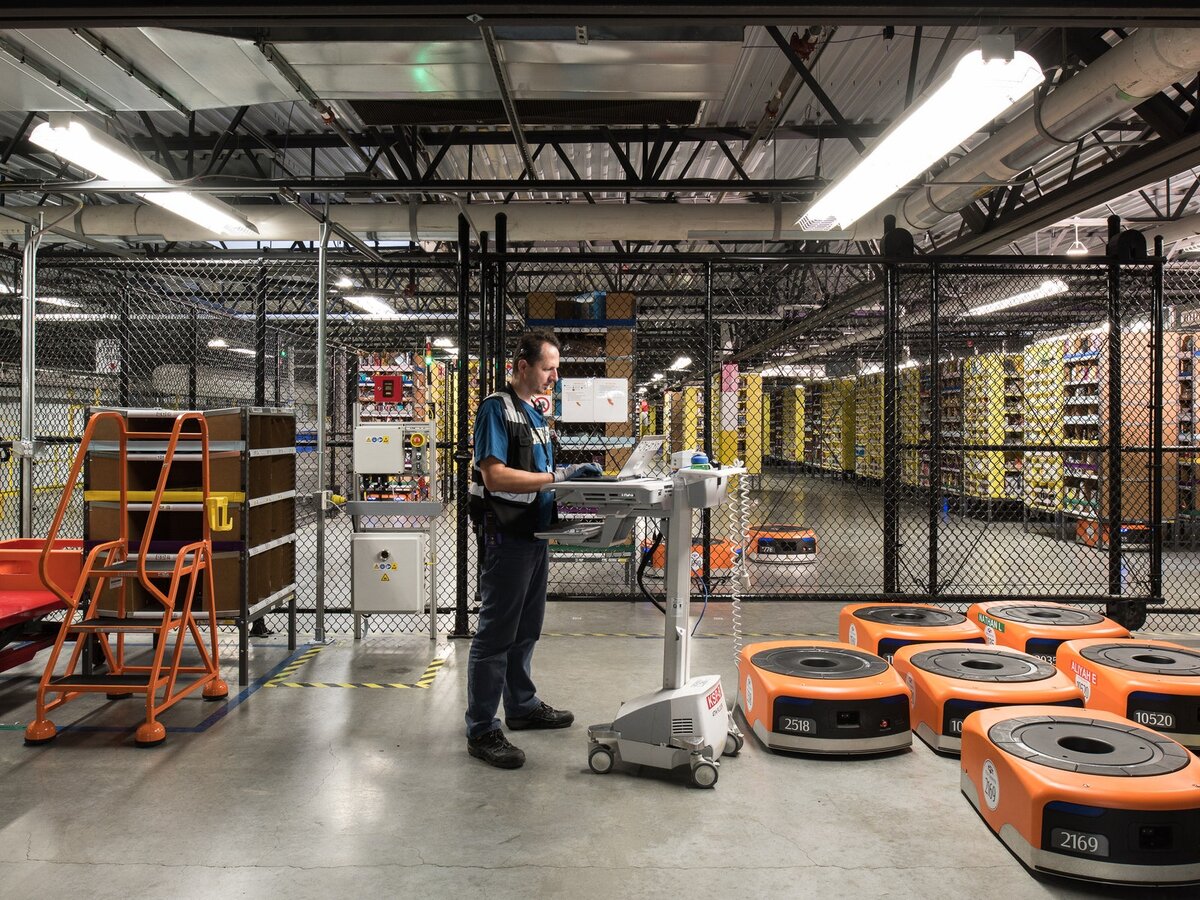 Техник Amazon работает на подставке для мобильного ноутбука. За ним клетка, где бродят складские роботы.