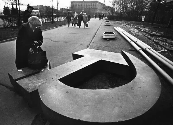Женщина что-то ищет в своей сумке., которую она поставила на упавший символ СССР. Москва, 1991 год