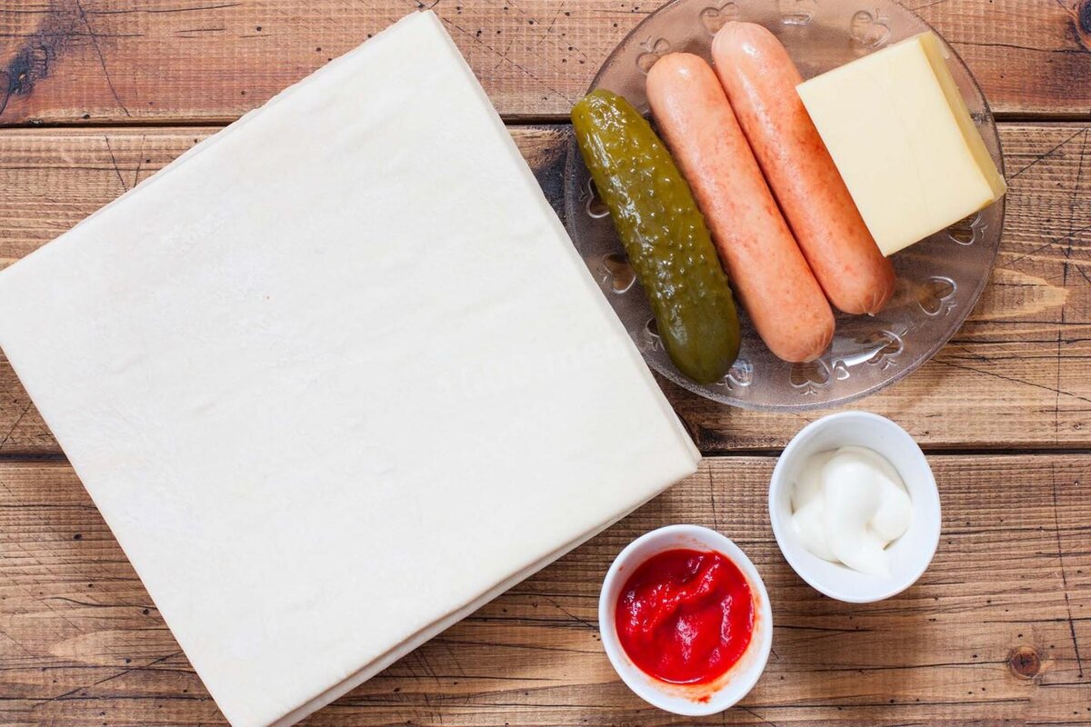 есто слоёное можно взять магазинное, можно приготовить самостоятельно. При выборе слоёного теста отдайте предпочтение бездрожжевому варианту, такое тесто лучше сохранит начинку и будет полезней для фигуры.  Сосиски выбирайте проверенного производителя с хорошим составом. Огурцы маринованные берите не кислые. Кетчуп и майонез можно заменить на свежие томаты.