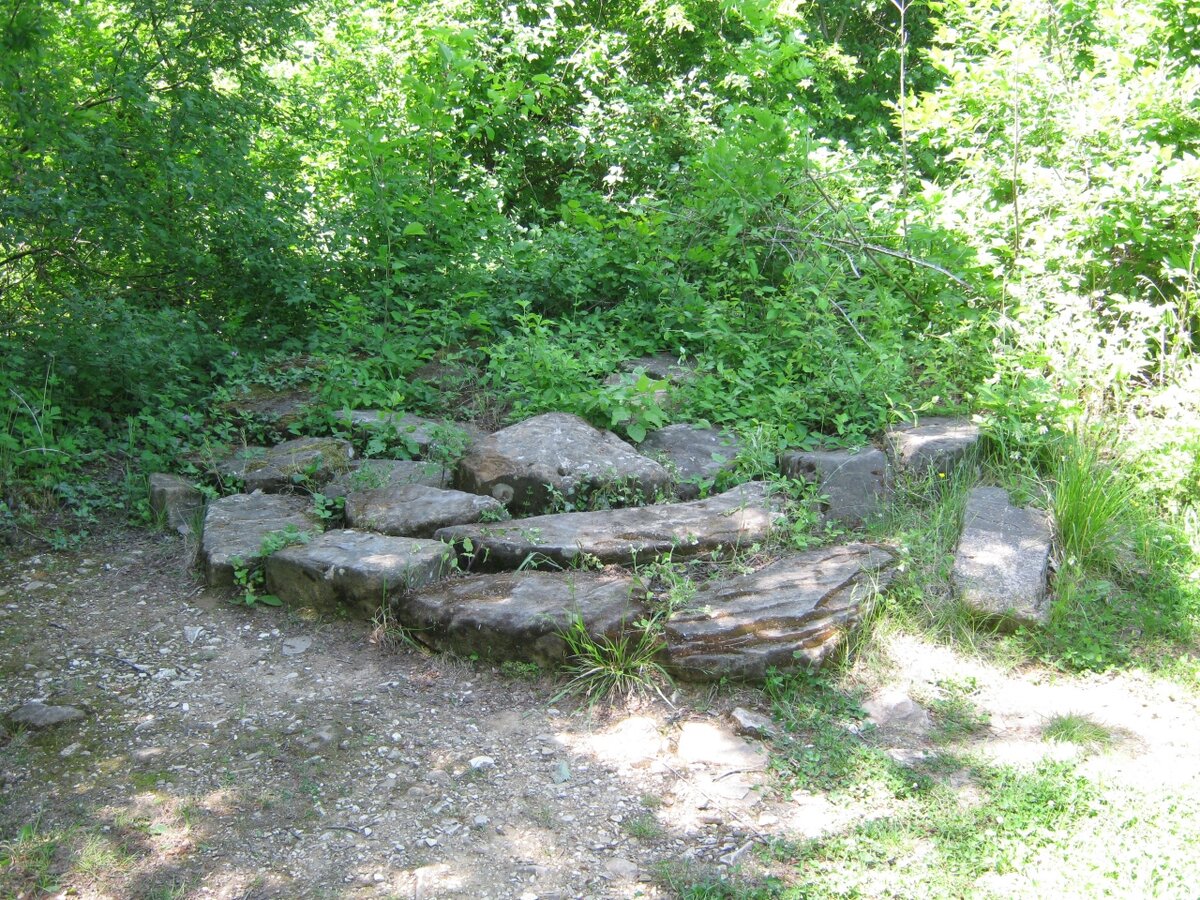 Находится между между дольменами Гуна и Саны, перед ними в 20 метрах Я вижу человека средних лет, высокого роста, с крупными чертами лица, белой кожей, серыми глазами, без бороды и усов. Волосы седые, белые, волнистые, до плеч.
Одет в тёмное платье до пят с белым воротничком, расклешённое книзу, подпоясан.