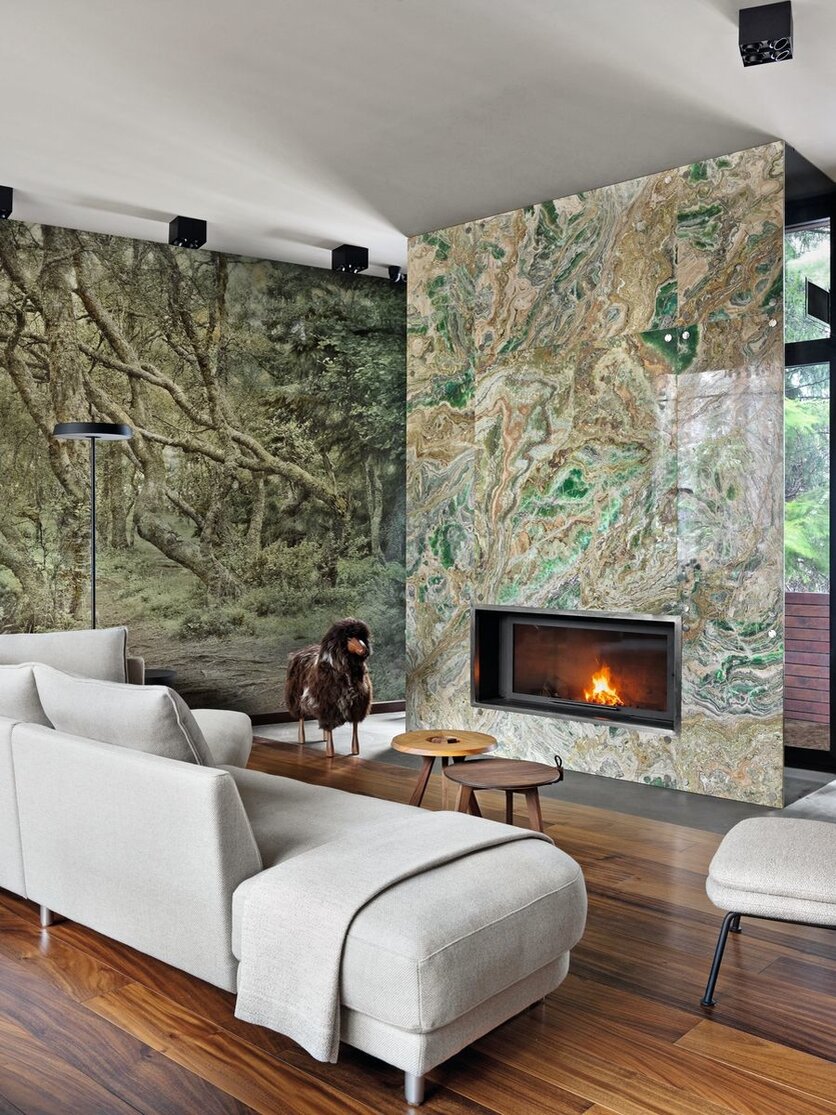 Главную гостиную украшает камин в обрамлении зелёного оникса. Большое панно из натурального камня вступает в диалог с зелёным массивом за окнами, а поддерживает тему природы в интерьере реалистичное изображение леса на одной из стен. Все системы освещения в доме лаконичны и решены в чёрном цвете. Светильники, Kreon. Диваны, Rolf Benz.