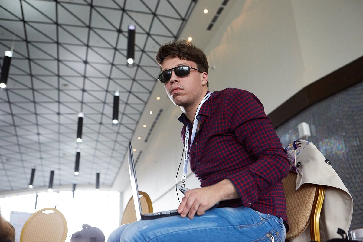 Тифлокомментарий. Фото в помещении. Ракурс немного снизу. Евгений Некрасов сидит на бежевом стуле. На коленях у мужчины стоит открытый ноутбук, Евгений придерживает его рукой. На молодом человеке темные очки, на длинной ленте бейдж. Евгений одет в красную клетчатую рубашку и синие джинсы. На заднем плане видно ещё несколько бежевых стульев. На потолке много небольших точечных чёрных светильников. 