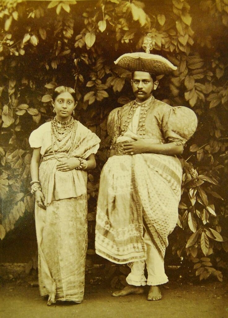 Придворный костюм кандийского Адикара — министра государства и юстиции — состоит из квадратной шапки, похожей на огромную подушечку для иголок. посередине увенчан драгоценным камнем. Куртка из ткани, с короткими плетеными рукавами, очень широкими в плечах и застегнута аметистовыми пуговицами, поверх нее надет белый палантин из плетеного муслина с золотым кантом. часть туловища, поверх белых штанов, обтягивающих щиколотку и оканчивающихся оборкой, ряд белых муслиновых и золотых узорчатых тканей перевязан громоздкими складками вокруг талии широким золотым поясом.У знатной жены местного сановника одежда больше сидит по фигуре, нет головного убора, а в волосах золотые украшения. Волосы длинные, гладко разделенные от середины лба и собраны в узел. Маленькие руки, увешанные многочисленными браслетами, завершают украшение кандийской аристократки. На фото министр с женой. Шри-​Ланка, (Цейлон). 1890-е годы.
