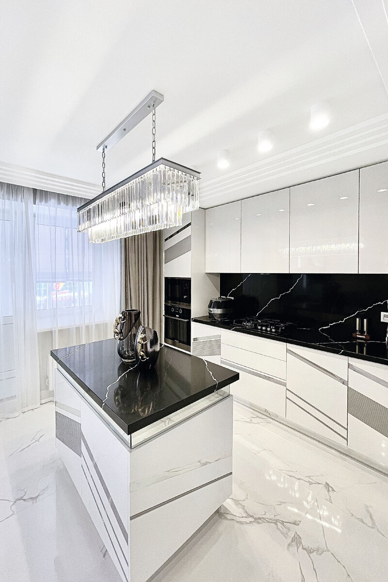 Белая плитка напольного покрытия и черные столешницы с фартуком на кухне прибавляют необходимый цветовой контраст интерьеру, а также гармонично перекликаются с панно и декоративными вставками в спальне. Строгий геометрический декор на мебели и в оформлении стен сглаживают мягкие узоры под мрамор на полу и поверхностях столов.