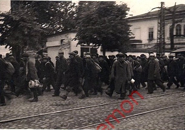 Идут по улице  Берхрессаденер штрассе (она же Великолукская, Советская). Идут мужчины в гражданском, немногие в форме. У многих в руках сумки, рюкзаки, скрутки и котелки. На первом плане видны рельсы. До войны и вовремя по городу ходили трамваи. Депо было тут рядом. За колонной будка,  совсем забор снесли - поставили кованный. До последнего времени ее использовали, как щитовую. Справа металлический столб. Справа усадьба Карамышева. Ныне разрушена. Листайте дальше локация. 