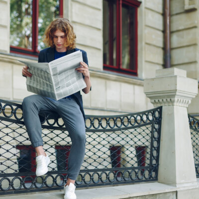    Young reddish, red haired attractive man with curly hair, reading a newspaper sitting near an urban old style building. Youth in action young leader getting to know world, city news. Валерий