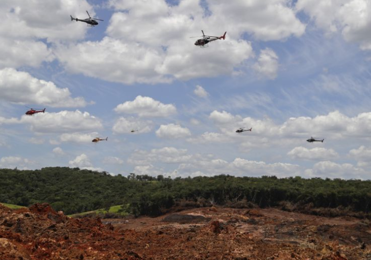 Вертолеты парят над горнорудным комплексом, чтобы освободить тысячи цветочных лепестков, отдавая дань уважения десяткам убитых и десяткам пропавших без вести после того, как шахтная Дамба рухнула.