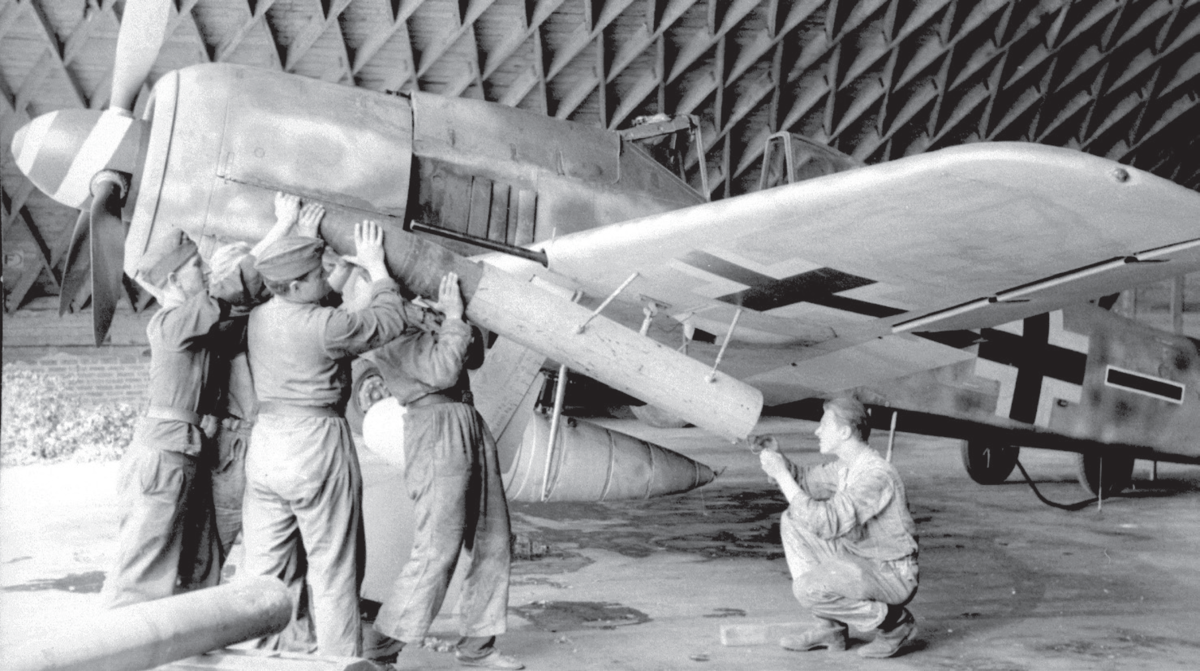 Авиабаза Лилль-Вандевиль, Франция, май 1944 г. — обычный  вид оружейников из Stab/JG 26, загружающих 21-см Werfer-Granate в пусковую трубу, подвешенную к левому крылу Fw 190A-8/R6, который также оснащен 300- литровым сбрасываемым баком для дальних полетов. Техник, присевший у задней части трубы, готов подключить провода зажигания и проверить резьбовой болт, который удерживает мину-ракеты. 