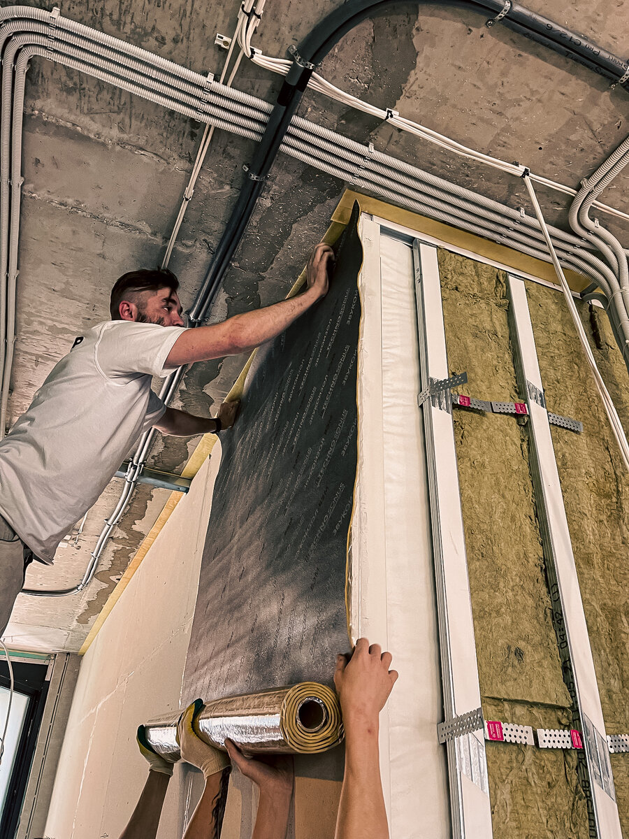 Мастера выполняют звукоизоляцию стены от соседей сбоку. Клеят мембрану на листы гипсокартона, чтобы сделать стену тяжелее! 