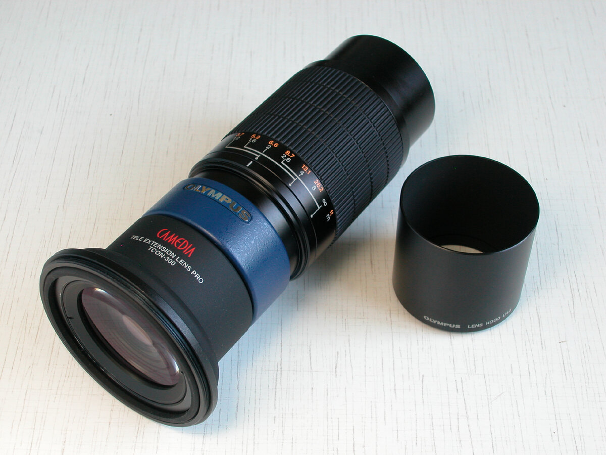 Olympus TCON-300. На фото изображён с комплектной металлической блендой. На резиновую муфту, поверх неё, надето переходное кольцо (необходимо для подсоединения Panasonic FZ50, не мешает установке Olympus E-20).