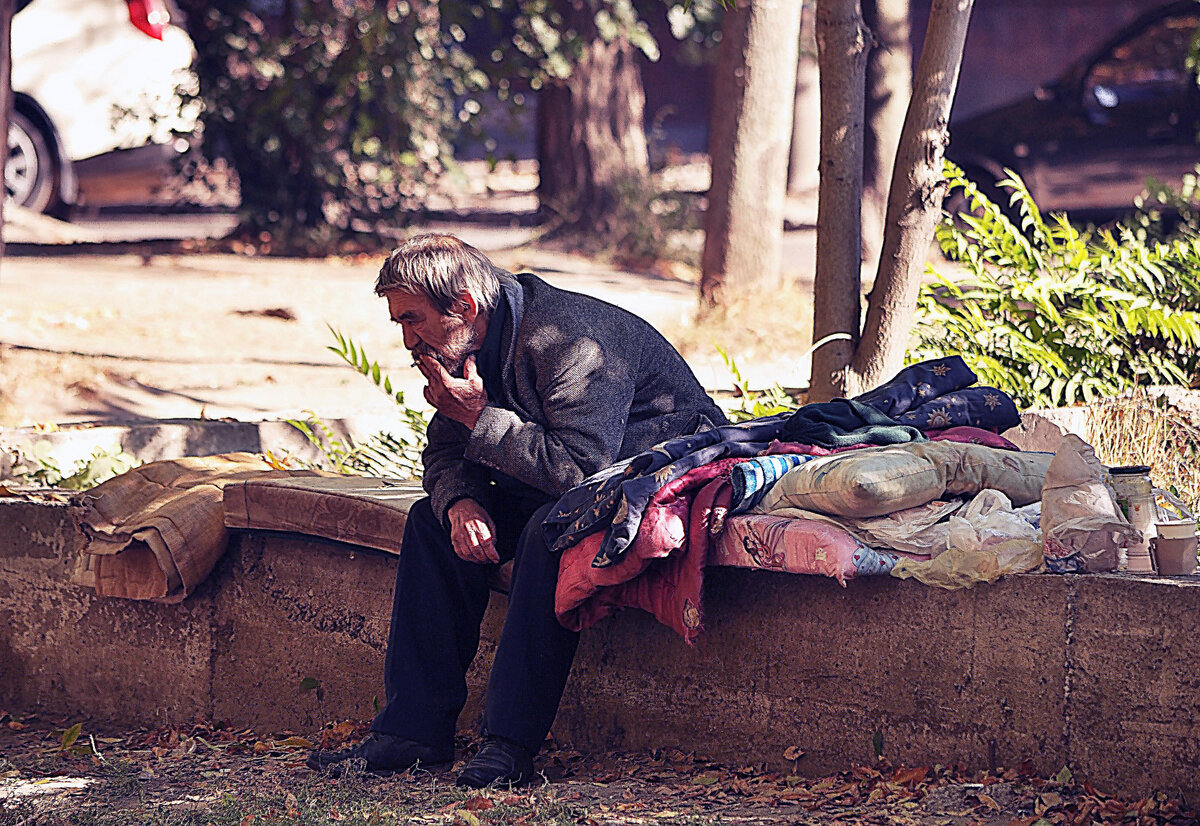 Бездомный курит сидя на матрасе.