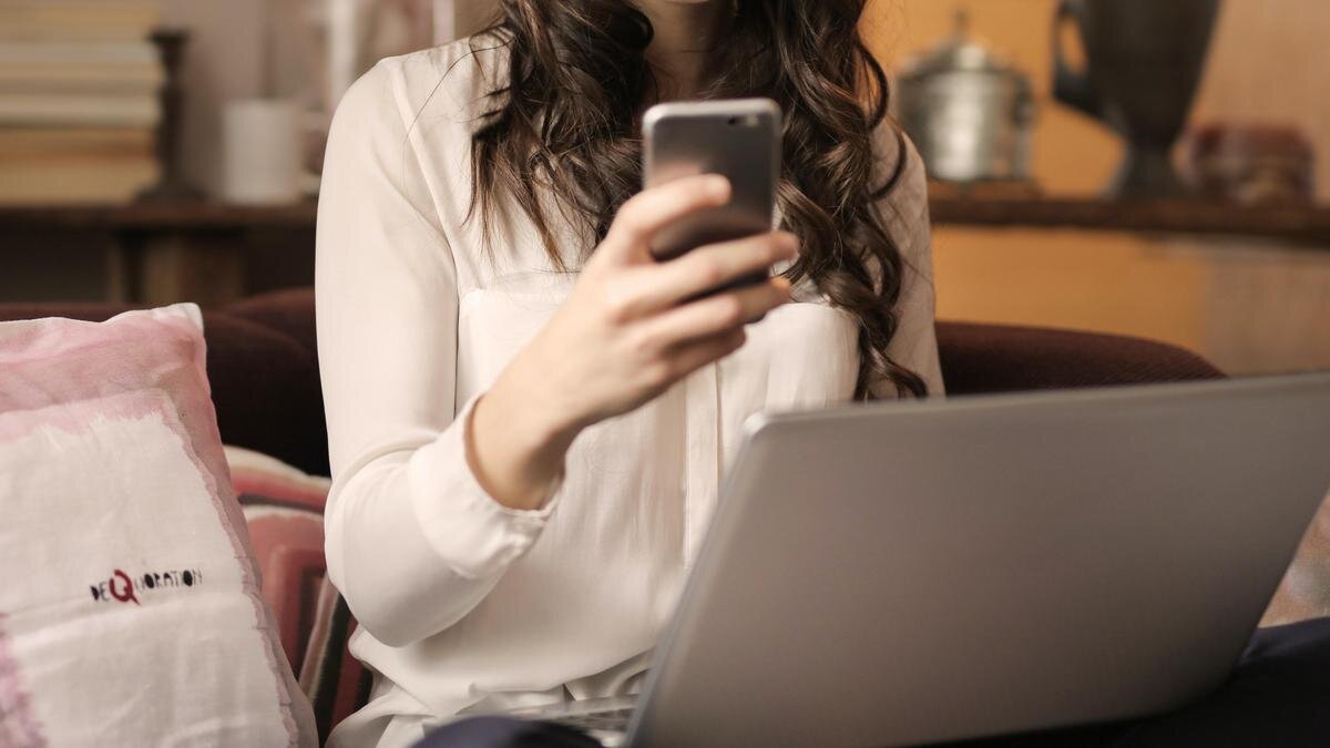    Девушка с ноутбуком на коленях улыбается, глядя в телефон pexels.com