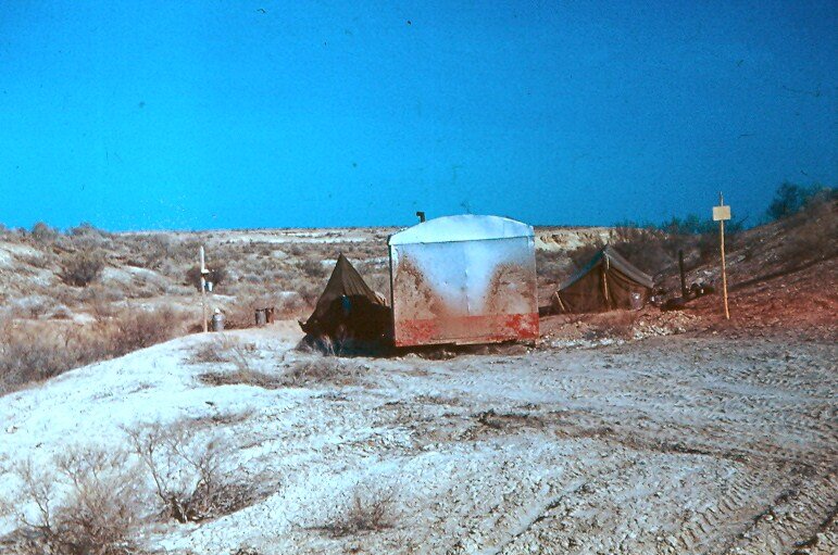 Справа от вагончика стоит первая палатка. Слева - бочка с водой, укрытая тентом от солнца. На табличке на шесте справа от палатки надпись "Капланкырский заповедник"