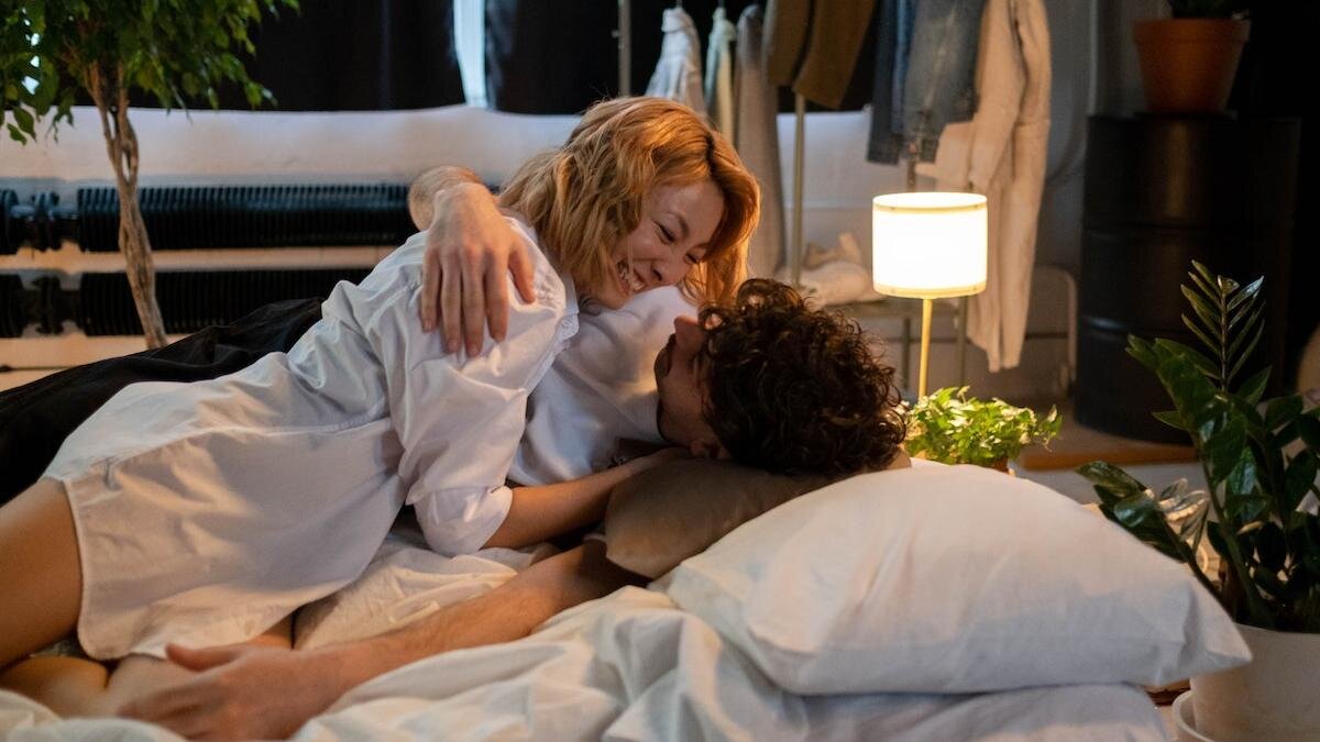    Влюбленная пара смеется и обнимается, лежа в постели на фоне окна:Pexels