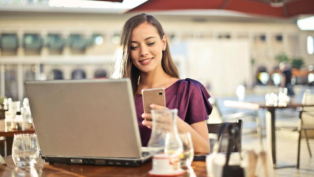    Девушка с телефоном в руках работает за ноутбуком в кафе:Pexels