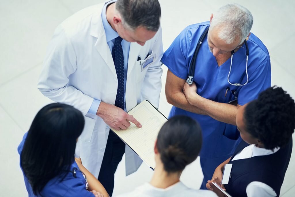    Shot of a group of doctors talking together over a medical chart while standing in a hospital GxP News