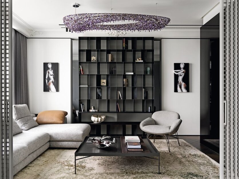 Доминантой в гостиной служит угловой диван Atlas, Arketipo. А оригинальным интерьерным элементом стал стеллаж из воронёной стали, выполненный по авторским чертежам. Его грани расположены в хаотичном порядке и развёрнуты к зрителю под разными углами, что позволяет конструкции выглядеть самодостаточной и без декоративного наполнения. В центре гостиной — объёмная люстра из цветных кристаллов богемского стекла Artica Bohemian от Manooi, кропотливо собранная и установленная мастерами компании HomeArt | Interior Showroom. Картины, Franc¸ois Sahuc, Carré d’Artistes. На стеллаже — керамика, Лиза Федотова, Lf Ceramics. Портьеры, компания «Придворный портной».