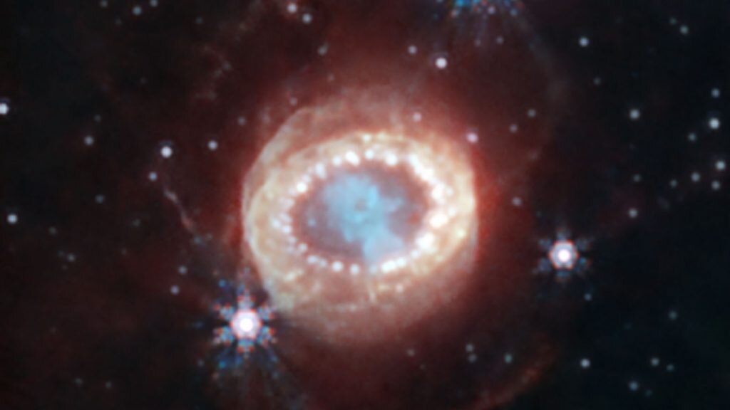    The NASA/ESA/CSA James Webb Space Telescope has begun the study of one of the most renowned supernovae, SN 1987A (Supernova 1987A). Located 168,000 light-years away in the Large Magellanic Cloud, SN 1987A has been a target of intense observations at wavelengths ranging from gamma rays to radio for nearly 40 years, since its discovery in February of 1987. New observations by Webb’s NIRCam (Near-Infrared Camera) provide a crucial clue to our understanding of how a supernova develops over time to shape its remnant. This image reveals a central structure like a keyhole. This center is packed with clumpy gas and dust ejected by the supernova explosion. The dust is so dense that even near-infrared light that Webb detects can’t penetrate it, shaping the dark “hole” in the keyhole. A bright, equatorial ring surrounds the inner keyhole, forming a band around the waist that connects two faint arms of hourglass-shaped outer rings. The equatorial ring, formed from material ejected tens of thousands of years before the supernova explosion, contains bright hot spots, which appeared as the supernova’s shock wave hit the ring. Now spots are found even exterior to the ring, with diffuse emission surrounding it. These are the locations of supernova shocks hitting more exterior material. In this image blue represents light at 1.5 microns (F150W), cyan 1.64 and 2.0 microns (F164N, F200W), yellow 3.23 microns (F323N), orange 4.05 microns (F405N), and red 4.44 microns (F444W). Александр Шереметьев