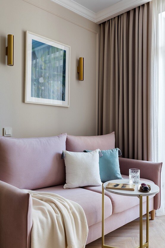 Розовый диван выделяется на фоне отделки, углубляя палитру интерьера. Умеренными акцентами стали голубые пятна — декоративная подушка и картина. Ещё гамму разнообразили с помощью элементов под латунь — опоры кофейного столика, подсвечников, перегородки, осветительных приборов.