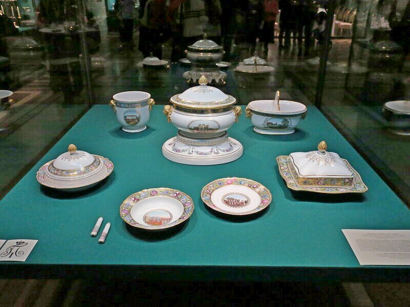 Предметы Кабинетского сервиза. 2016-й, декабрь. Фото с выставки "Убранство русского императорского стола XVIII - начала XX века". Видим супницу на великолепной фарфоровой подставке, маслёнку с крышкой, ещё одно блюдо с крышкой, название которого, надеюсь, подскажут знатоки, тарелку под первое и подтарелочник, а также нечто, похожее на вазу с хватами-маскаронами, и менажницу(?) под закуски\салаты. Фото взято из свободного доступа в интернете.