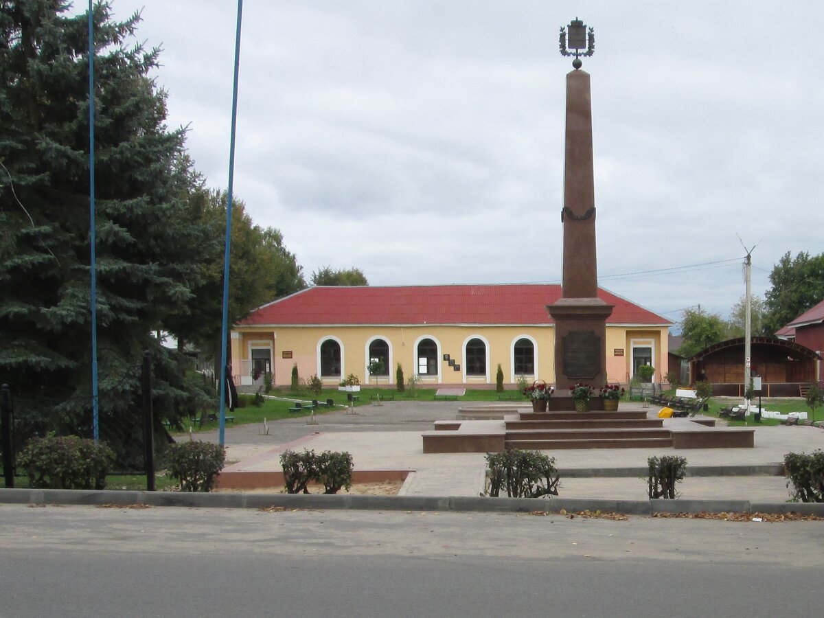 Стела, установленная в честь присвоения Жиздре звания "Город воинской доблести". на заднем плане -- районная библиотека с памятными досками, посвящёнными всемирноизвестному учёному-генетику Н.П. Дубинину.