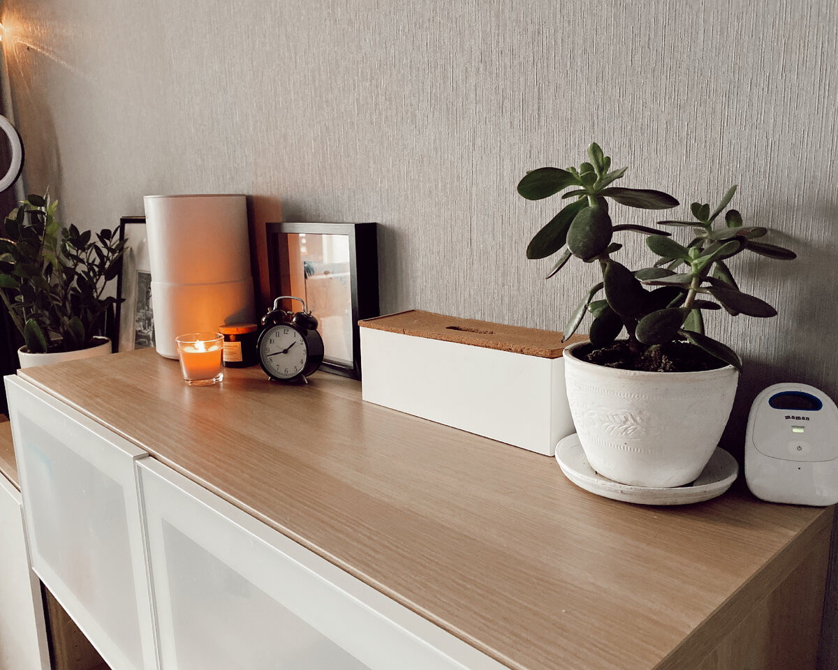 На своем импровизированном комоде храню как необходимые - радионяня, увлажнитель воздуха, часы, короб с проводами и розетками (пилот), так и вещи для души. Избавилась от искусственных цветов. Хочу, чтобы в моем доме все было максимально натурально. Из декора живые цветы, подсвечник из соли, который благотворной влияет на воздух, друзы кристаллов (подарок мамы для моей коллекции), аромасвечи и картины. Разноцветные горшки я покрасила в белый цвет, чтобы они подходили по стилю.