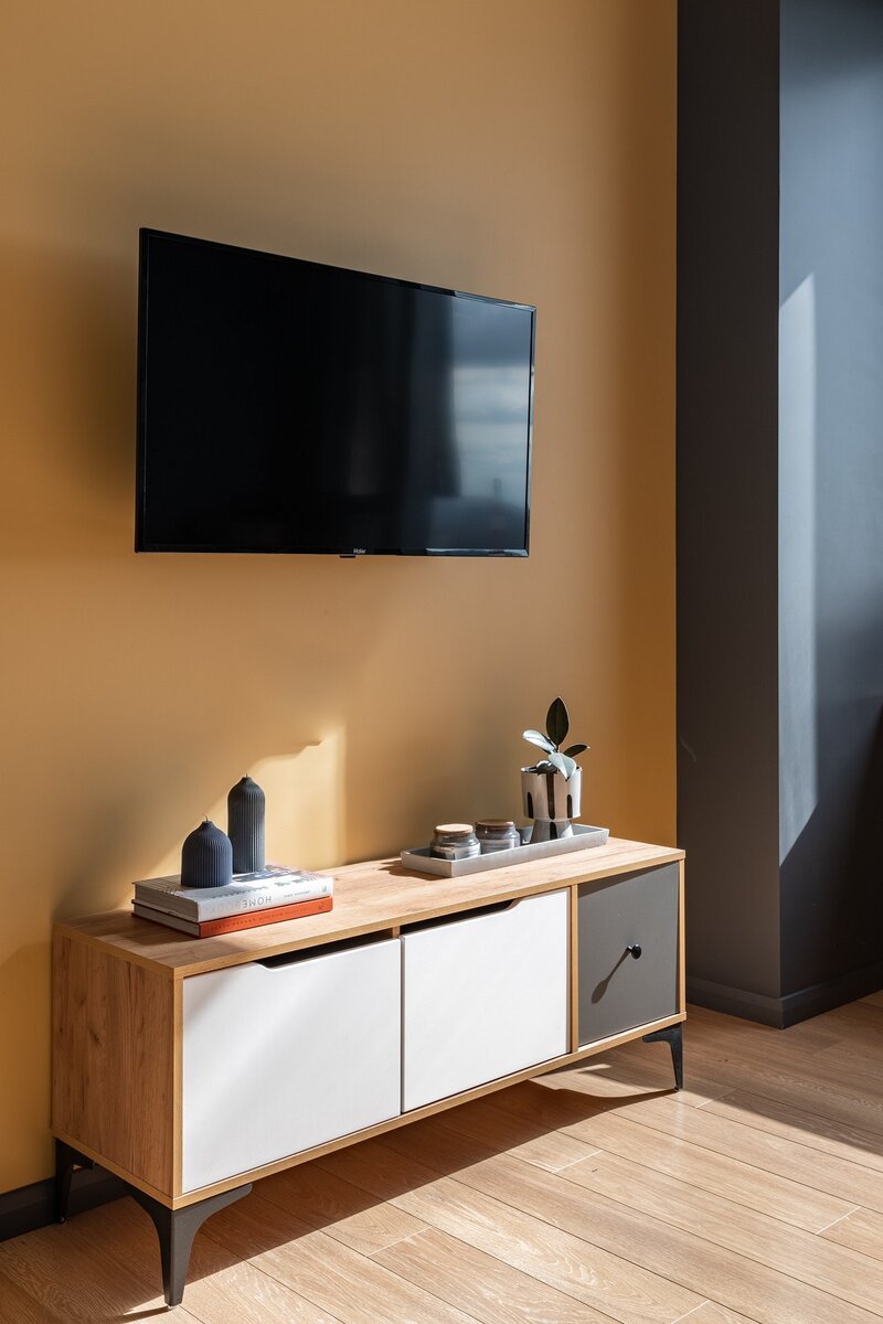 Напротив оборудовали ТВ-зону. Стену выкрасили в горчичный цвет. Оттенок подбирали по обоям за изголовьем. Такой подход помог собрать детали проекта в единую концепцию.