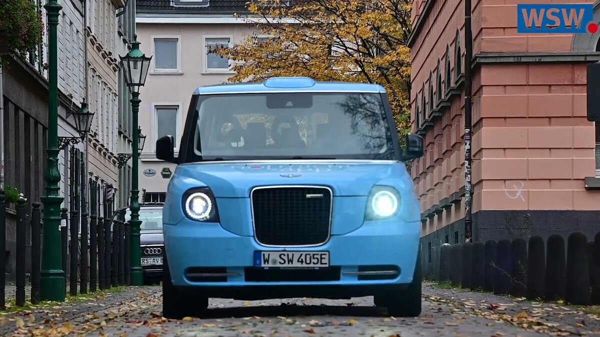 Светло-голубой электромобиль модели LEVC TX проезжает по жилому району в районе Эльберфельд.