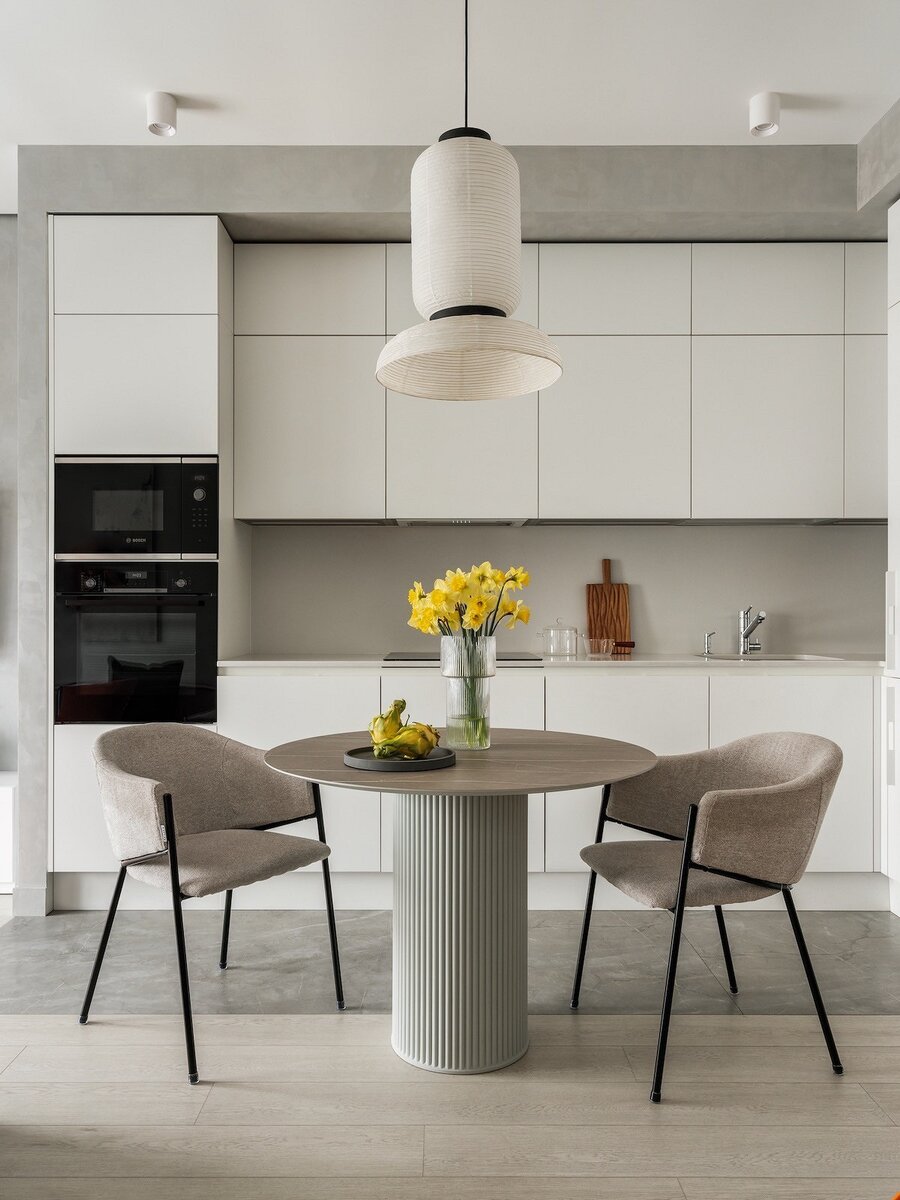 Угловой гарнитур встроили в нишу. Ряды шкафов обрамили высокими пеналами для техники по краям. Белые фасады помогли визуально уменьшить роль конструкции в интерьере. Предусмотрели не только верхний ярус, но и антресольный ряд, что позволило вместить всё необходимое. При этом за счет спокойной палитры кухня не кажется слишком громоздкой. Листайте галерею →