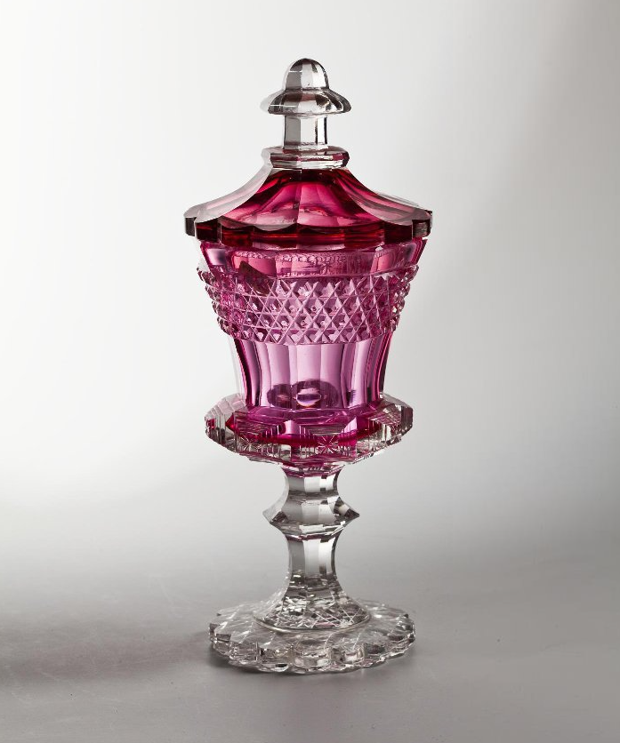 Императорский стеклянный завод. Кубок с крышкой. Российская империя, г. Санкт-Петербург. Вторая четверть XIX века. Малиновый кубок, декорированный полосой мелкой ромбической грани в верхней части («strawberry diamond»), выполнен в стиле модных во второй четверти XIX века готических реминисценций. Об этом свидетельствует его форма, напоминающая средневековые кубки «с кровлями», четкое и ритмичное зональное разграничение элементов виртуозной декоративной грани, яркий колористический контраст чистейшего алого и бесцветного стекла. Подобные кубки носили, чаще всего, подарочный, репрезентативный характер и относились к ассортименту дорогих изделий ИСЗ.