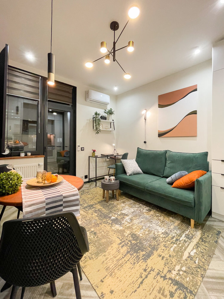 В роли цветного акцента выступает диван. Его можно разложить, если гости останутся на ночь. Рядом поставили мягкий пуф-табурет, который взял на себя функцию журнального столика. В шкаф рядом с диваном вмонтировали розетки, чтобы убрать туда чайник и кофемашину.