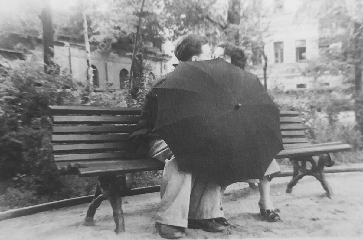 Молодая пара на скамеечке прикрывается зонтом, чтобы поцеловаться.