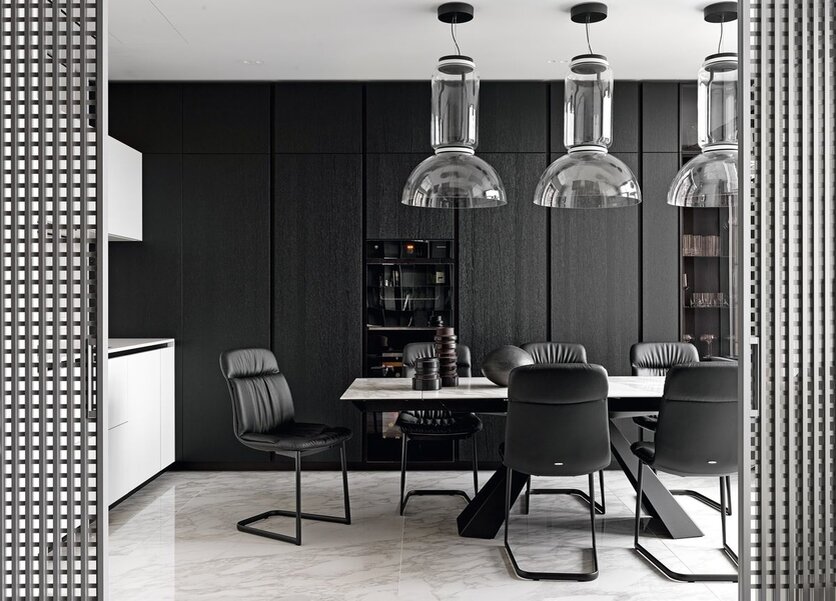 Кухня оборудована по последнему слову техники — от духового шкафа с системой подогрева паром до лёдогенератора в холодильнике, всё — от компании V-ZUG. На фоне тёмной кухни выразительно смотрятся обеденная группа Cattelan Italia на шесть персон и три крупных прозрачных светильника Noctambule от Flos.