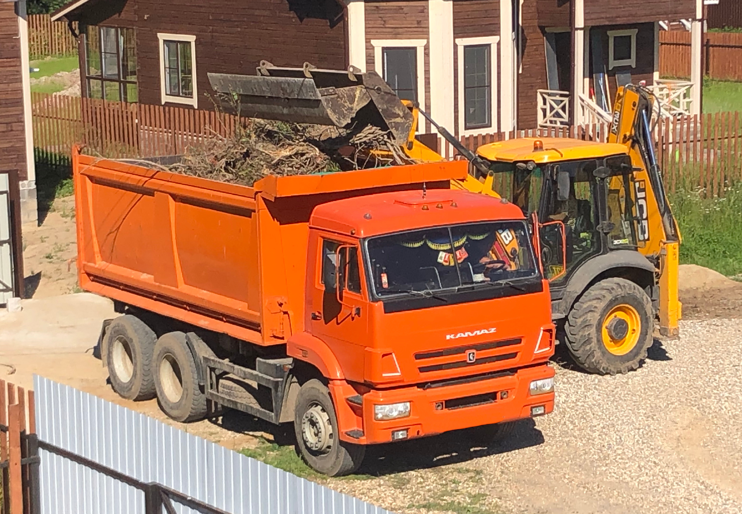 Экскаватор-погрузчик JCB загружает корни на самосвал КАМАЗ. Вывоз мусора с  дачного участка.