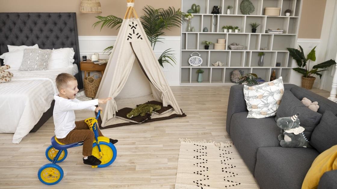    Мальчик ездит на трехколесном велосипеде по комнате, где стоит большая кровать, диван с подушками, вигвам, большая пальма. Возле стены размещен комод с открытми полочками, на которых стоят сувениры и комнатные растения:Freepik