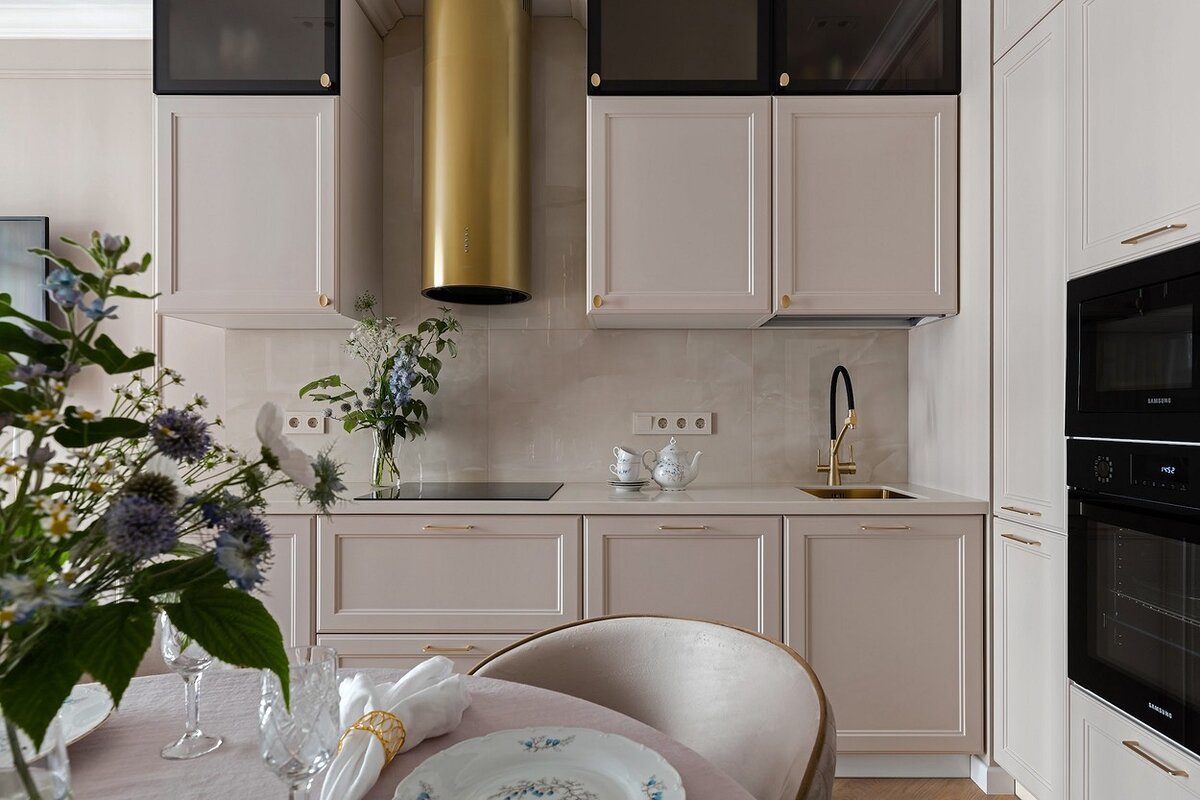 Чтобы интегрировать кухню в жилое помещение, фасады гарнитура подобрали в цвет стенам. Дверцы с филёнкой повторяют рисунок молдингов. Шкафы-пеналы и часть техники упаковали в нишу. Таким образом, вся зона выглядит очень лаконично. Выделяется только латунная вытяжка, но она хорошо перекликается с другими элементами в пространстве.