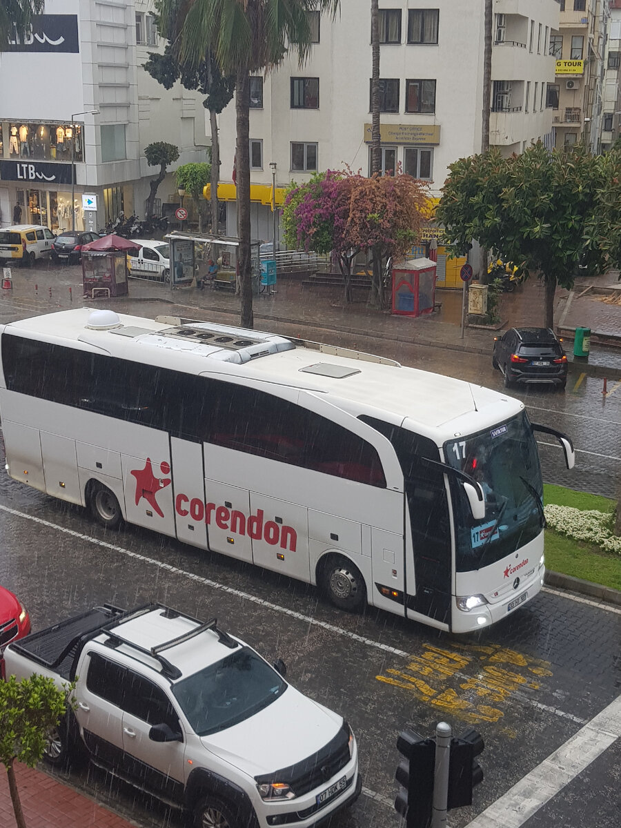 В редкий дождливый июньский день трансферный автобус  а/к "Corendon" везёт пассажиров на рейс, возможно, в Россию. (Ведь эта иностранная авиакомпания осуществляет перелёты из Анталии в РФ). На заднем плане фотографии: дворник прячется от дождя на автобусной остановке в Алании.