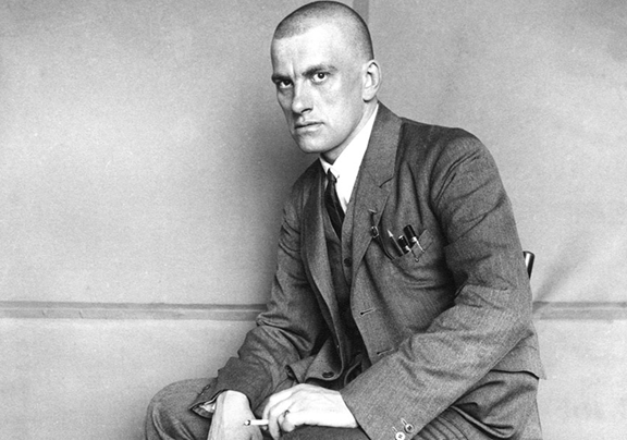 Владимир Маяковский сидит на стуле и курит, держа в правой руке шляпу, а в левой сигарету.