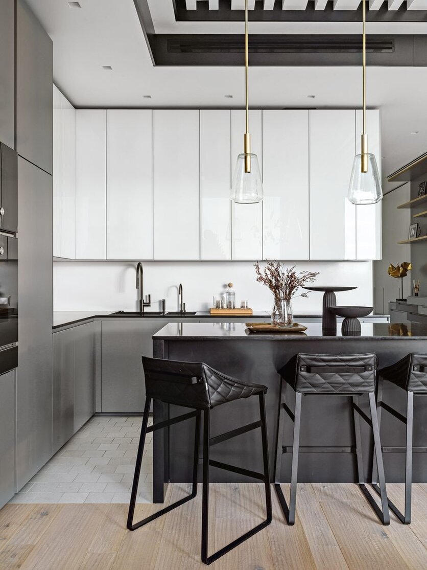Чтобы визуально объединить зону кухни и гостиной, стены в гостиной отделали панелями белого цвета с контрастным вертикальным швом. Аналогичным образом дизайнеры поступили на кухне — за белыми фасадами верхних шкафов оборудованы холодильник, винный холодильник, морозильник, вакууматор, пароварка и духовой шкаф.
